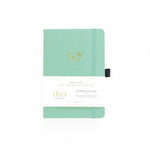 An image of a mint dot grid bullet journal with a gold foil mint envelope and pen on the cover. The journal is on a white background