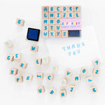 A flat image of wooden alphabet stamps and a blue ink pad. Blank white card features the words "Thank You" stamped on them using the wood stamps and ink pad. 