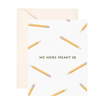 A folded greeting card with text that reads "We were meant to 2B" with cute illustrated pencils. Card is lying on a light pink envelope.