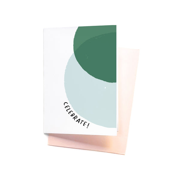 A folded greeting card with text that reads "Celebrate!" with an illustration of overlapping circles. The card is lying on a blush pink envelope.