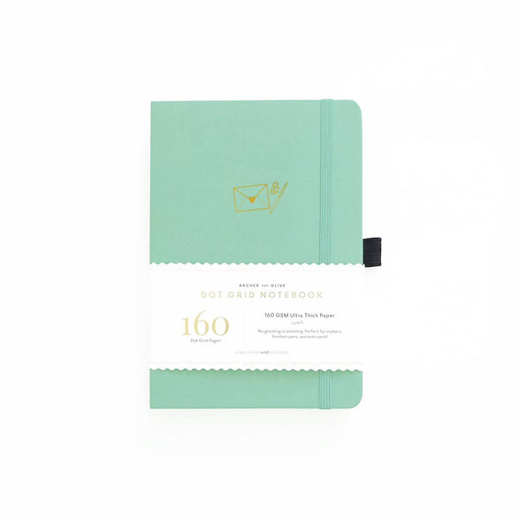 An image of a mint dot grid bullet journal with a gold foil mint envelope and pen on the cover. The journal is on a white background