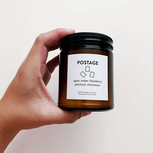 An image of a hand holding a small Paper + Craft Pantry Postage candle. The label says "Postage; apple, amber, blackberry, patchouli, chartreuse; Made with vegan coconut wax. Hand poured in Austin, Texas" with a little trio of three handdrawn postage stamps.