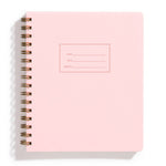 An image of a light pink notebook with gold spiral binding on a white background. The cover of the notebook has a small section to label the name, date and subject.
