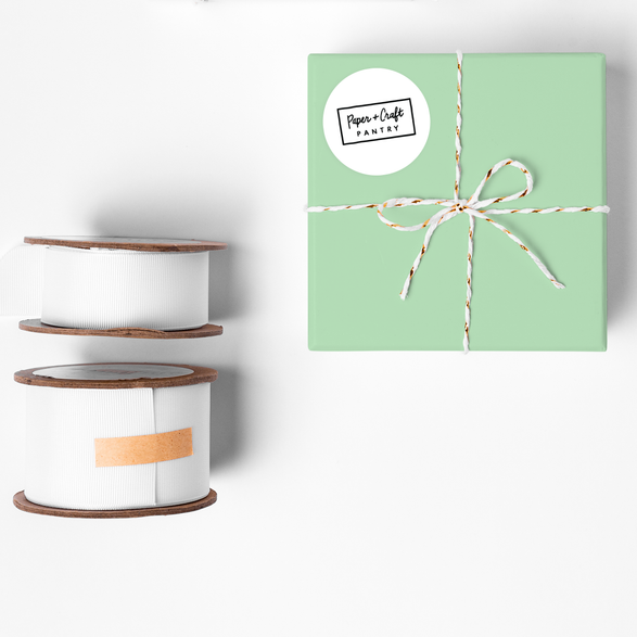 A gift with mint wrapping paper next to ribbon spools. The gift has a white round sticker label with the Paper + Craft Pantry logo on it.