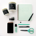 An image of three different size Paper Shop candles with assorted stationery around it. There is a mint notebook, a black kaweco fountain pen, a mini black notebook, mint stabilo highlighter, my favorite pen, green dual tipped marker, black emott pen, and a pair of mint scissors